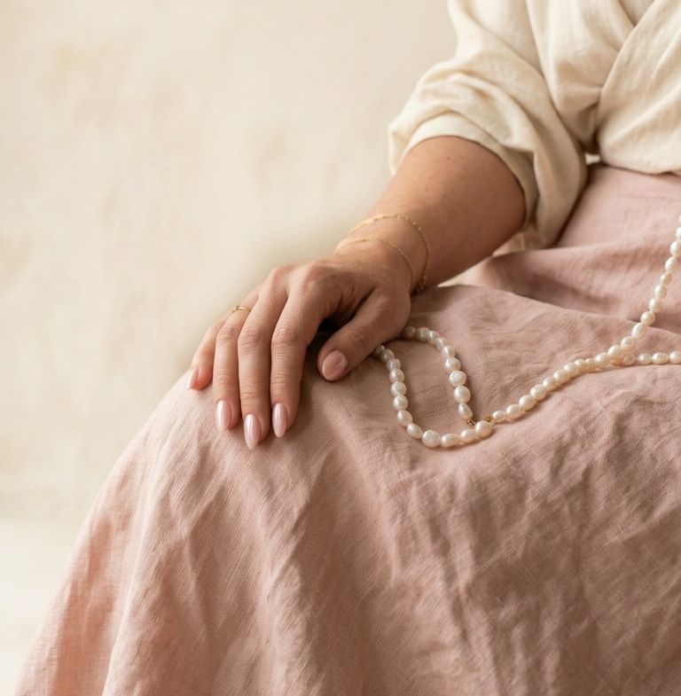 A woman seated in cream and blush linen, her hand resting on her knee beside a long strand of pearls.